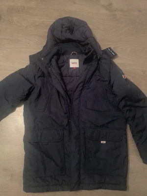 Mörkblå parkas från Tommy Jeans - Snygg mörkblå parkas från Tommy Jeans med huva och flera praktiska fickor framtill. Jackan har dragkedja och tryckknappar, samt en liten logotyp på ärmen. Perfekt för kalla dagar och riktigt stilren design.
