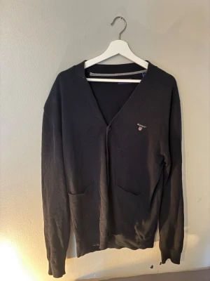 Svart cardigan från Gant - Svart cardigan från Gant med V-ringning och två fickor framtill. Diskret Gant-logga broderad på bröstet. Klassisk och stilren design, perfekt att slänga över en t-shirt. Mjukt material som känns skönt mot huden. Passar som M. Hör av er vid frågor - vid snabba köp kan priset sänkas🔥