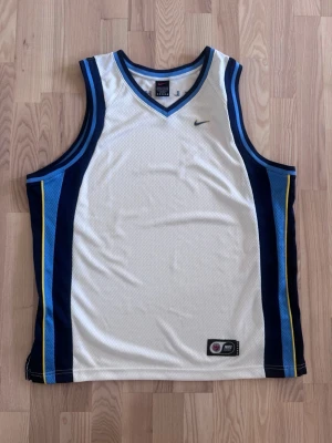 Nike basketlinne vitt och blått - Snyggt basketlinne från Nike i vitt med blå och ljusblå paneler samt gula detaljer. Modellen har V-ringning och ärmlös design, tillverkat i meshmaterial för bra ventilation. Ordet 'FLIGHT' står på ryggen och Nike-loggan finns på bröstet.