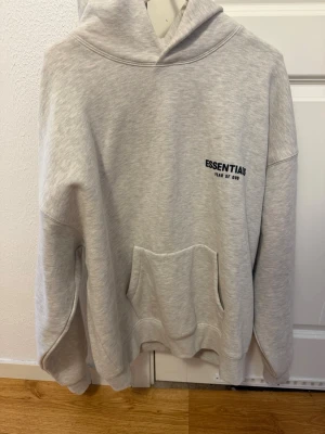 Grå Essentials Fear of God hoodie - Säljer en ljusgrå hoodie från Essentials Fear of God med svart logga på bröstet och stor text på ryggen. Hoodien har en stor känguruficka framtill, huva och ribbade muddar. Perfekt för dig som gillar streetwear och vill ha en clean look. Den är helt ny oanvänd utan prislapp dock. Vid intresse hör av dig! Priset kan diskuteras närmare vid intresse.
