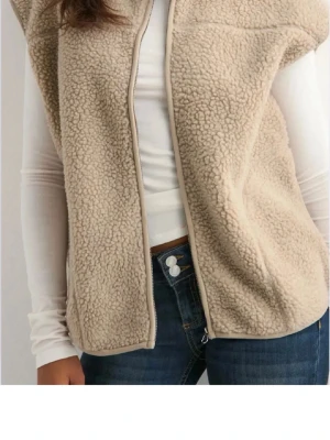 Beige teddyväst med dragkedja - Mysig beige teddyväst med hög krage och dragkedja framtill. Västen har en avslappnad passform och är gjord i mjukt, fluffigt material som ger en trendig look. Från Nelly