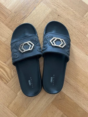 Svarta tofflor från ASOS DESIGN - Säljer ett par svarta tofflor från ASOS DESIGN med präglat blommönster och guldfärgade hexagonala detaljer på remmen. Tofflorna har en klassisk slip-in modell och är tillverkade i syntetmaterial. Perfekta för en chill och trendig look.