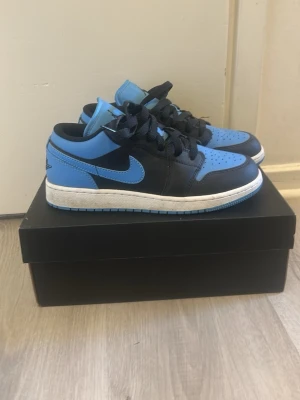 Air Jordan 1 Low blå och svart - Snygga Air Jordan 1 Low sneakers i blått och svart läder med klassisk svart swoosh och svarta skosnören. Skorna har perforerad tåbox och vit mellansula med blå yttersula. Perfekta för dig som gillar streetwear och vill sticka ut med en ikonisk modell.