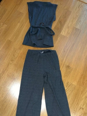 Mörkgrått ribbat set med bälte - Tvådelat set i mörkgrå ribbad trikå med ärmlös topp och matchande byxor. Toppen har ett knytbälte i midjan och rund halsringning. Materialet är mjukt och stretchigt, perfekt för en avslappnad men snygg look.