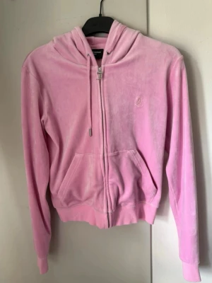 Rosa velourhoodie Juicy Couture - Mjuk och lyxig rosa velourhoodie från Juicy Couture med dragkedja, huva och fickor framtill. Ikoniskt JC-märke på bröstet och snygga detaljer på snörena. Perfekt för en avslappnad och trendig look.
