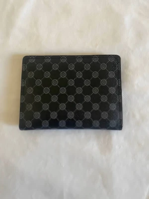 Svart plånbok med monogrammönster - Snygg svart plånbok med grått monogrammönster över hela ytan. Plånboken har flera kortfack, ett fönsterfack för ID och stängs med tryckknapp. Tillverkad i ett glansigt konstlädermaterial som känns både modernt och stilrent.