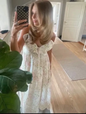 Blommig långklänning med volang - Söt långklänning med blommigt mönster i ljusa färger. Klänningen har volangdetaljer vid axlarna och en v-ringad urringning med rosett framtill. Perfekt för sommarens alla tillfällen.