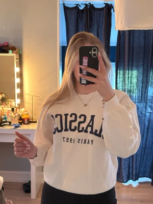 Vit sweatshirt med blå text - Säljer en vit sweatshirt med trycket 'CLASSICS PARIS 1997' i mörkblått framtill. Tröjan har rund halsringning, ribbade muddar vid ärmslut och nederkant samt en avslappnad passform. Perfekt för en chill och stilren look.