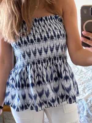 Blåvit mönstrad peplum topp - Säljer en peplum topp i blått och vitt med ett snyggt abstrakt mönster. Toppen är axelbandslös och har smock upptill för en bekväm passform samt en lätt volang nedtill. Från gina tricot! Passar S och M! Nästan aldrig använd. Inga fläckar eller så!🌸