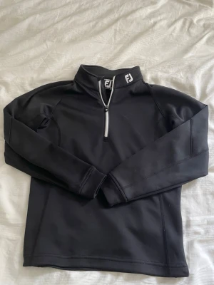 Svart sporttröja från FootJoy - Svart långärmad sporttröja från FootJoy med halv dragkedja framtill och hög krage med vit logga. Tillverkad i ett stretchigt funktionsmaterial som passar perfekt för träning eller golf. Snygg och stilren design med diskreta sömmar.