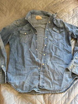 Ljusblå jeansskjorta med tryckknappar - Säljer en klassisk ljusblå jeansskjorta med långa ärmar och två bröstfickor(äkta). Skjortan har vita tryckknappar framtill och vid ärmsluten. Skjortan är från polo Ralph Lauren och är i perfekt skick! 