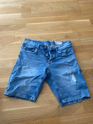 Blå jeansshorts med slitning - Säljer ett par blå jeansshorts med knappar i gylfen och slitning på ena benet. Shortsen har klassiska fem fickor och bälteshällor. Perfekta för varma dagar och ger en avslappnad look.