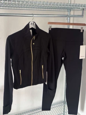 Svart träningsset med jacka och tights - Snyggt svart träningsset med en figurnära jacka med guldiga dragkedjor och högt krage samt matchande tights med hög midja. Perfekt för gymmet eller löprundan. Materialet är stretchigt och följsamt för maximal komfort. Är tillgänglig i alla storlekar och byxan/ koftan kommer tillsammans !!