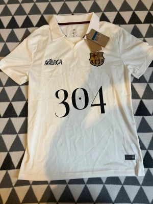 Vit FC Barcelona pikétröja - Säljer en vit pikétröja med FC Barcelona-emblem på bröstet och stor svart siffra 304 framtill. Tröjan har klassisk krage, korta ärmar och detaljer i svart och guld. Perfekt för dig som gillar fotboll och vill ha en stilren supportertröja.