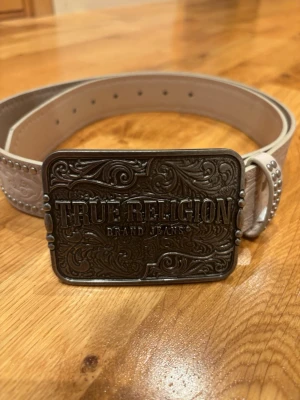 Vitt skärp från True Religion - Snyggt vitt skärp från True Religion med stor, detaljerad metallspänne och nitar längs kanten. Skärpet har en rektangulär buckle med ingraverad logga och mönster. Perfekt för att lyfta din outfit med en cool accessoar.