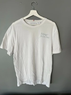 Vit t-shirt från Les Deux med tryck - Vit t-shirt från Les Deux med diskret text på bröstet och ett stort konstnärligt tryck på ryggen. Klassisk passform och rund halsringning. Perfekt för dig som gillar stilrena plagg med en twist.