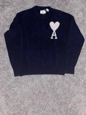 A - Mysig mörkblå stickad tröja med rund halsringning och ribbade muddar. På bröstet finns en vit hjärtdetalj med bokstaven A. Perfekt för kyligare dagar och enkel att matcha till olika outfits.