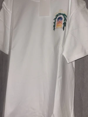 Vit t-shirt med tryck Avenida Casablanca - Vit t-shirt i bomull med färgglatt tryck av en byggnad och texten 'Avenida Casablanca' på bröstet och ryggen. T-shirten har rund hals och korta ärmar. Perfekt för dig som gillar unika motiv och stilrena plagg.