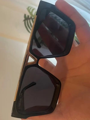 L*V solglasögon - Snygga svarta solglasögon från L*V med breda bågar och mörka glas. Ramen har en cool geometrisk form och detaljer i guld längs ovansidan. Perfekt accessoar för dig som vill sticka ut med en exklusiv look.
