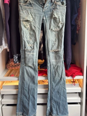 Ljusblå slitna bootcut jeans - Säljer ett par ljusblå jeans med bootcut passform och flera slitningar och hål på både fram- och baksida. Jeansen har klassisk femficksdesign och stängs med knappgylf. Perfekta för en avslappnad och trendig look. Finns hål på baksidan men är lätt att laga. Kan även tänka mig laga innan man köper men kommer tyvärr kosta extra, ungefär 150kr. De är bootcut men ser lite raka ut.
