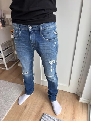 Replay Anbass Jeans  -  W30 L32 Säljer ett par blå jeans med slitningar och detaljerade revor på framsidan. Jeansen har fem fickor, normal midja och raka ben. Materialet är klassisk denim och färgen är mellanblå med ljusare tvätt på vissa partier.