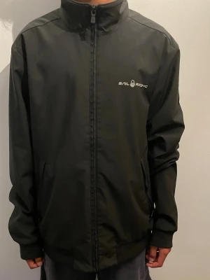 Svart softshell från Sail Racing - Stilren svart softshell jacka från Sail Racing med hög krage och dragkedja framtill. Jackan har logga på bröstet och en patch på ärmen. Tillverkad i ett lätt och tåligt material, perfekt för blåsiga dagar. Normal passform och långa ärmar.