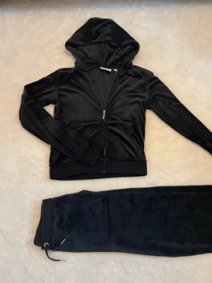 Svart velourset från Juicy Couture - Mysigt marinblått set från Juicy Couture med hoodie och dragkedja samt matchande byxor. Setet har klassisk Juicy Couture-detalj på snöret och är perfekt för chill dagar hemma. Settet är i storlek 12-13 år.