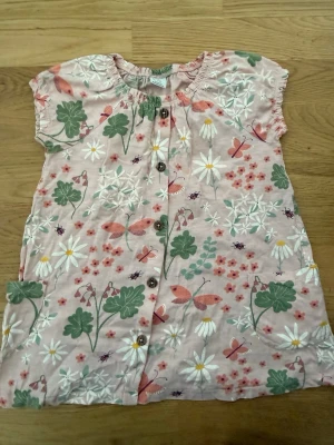 Blommig kort klänning från Lindex - Söt kort klänning från Lindex i storlek 104. Klänningen är ljusrosa med blommigt mönster i grönt, vitt och rosa samt fjärilar. Knappar framtill och två fickor. Perfekt för sommardagar!