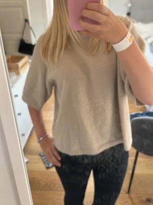 Beige stickad tröja med halvärm - Mysig beige stickad tröja med avslappnad passform och halvärmar. Tröjan har ribbade kanter vid hals, ärmslut och nederkant. Perfekt för lager på lager och enkel att styla till olika outfits.