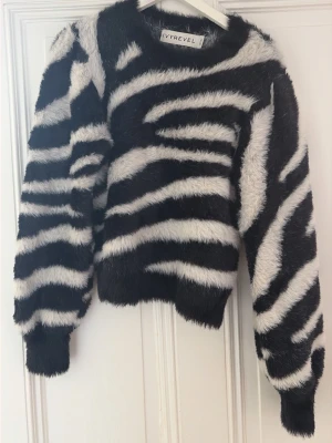 Zebramönstrad stickad tröja Ivyrevel - Säljer en fluffig stickad tröja från Ivyrevel med svartvitt zebramönster. Tröjan har rund hals, långa ärmar och en croppad passform. Materialet är mjukt och lurvigt, perfekt för dig som vill sticka ut med en djurinspirerad look.