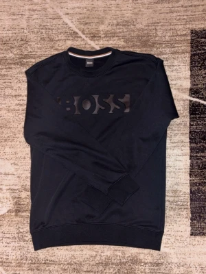 Svart sweatshirt från BOSS - Säljer en svart sweatshirt från BOSS i storlek M. Tröjan har ett stilrent BOSS-tryck framtill, rund halsringning och långa ärmar. Tillverkad i mjuk bomull för skön känsla och passar perfekt till en avslappnad look.