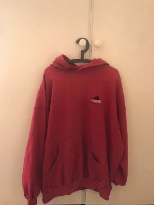Röd hoodie från Adidas Equipment 90 talet - Säljer en röd hoodie från Adidas Equipment med broderad logga på bröstet och en svart Adidas-patch på ärmen. Hoodien har stor luva, känguruficka och ribbade muddar. Perfekt för dig som gillar sportig och avslappnad stil.