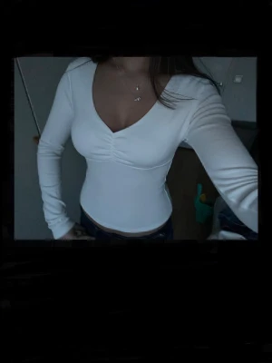 Vit långärmad topp med v-ringning - Säljer en stilren vit topp med lång ärm och snygg v-ringning. Toppen har en lätt rynkning vid bysten och sitter figurnära för en smickrande siluett. Perfekt att matcha med jeans eller kjol för en trendig look.