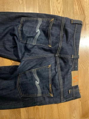 Mörkblå jeans från Nudie Jeans - Snygga mörkblå jeans från Nudie Jeans med klassisk femficksmodell och kontrastsömmar. Bakfickorna har den ikoniska vågiga sömmen och ett brunt läderpatch med logga i midjan. Jeansen har rak passform och är tillverkade i kraftig denim.