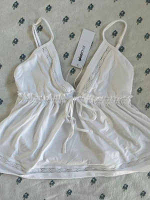 BNWT vit babydoll linnetopp från Gina Tricot - Söt helt ny vit babydoll linnetopp från Gina Tricot med tunna axelband, spetsdetaljer längs ringningen och nederkanten samt en dekorativ knytning i midjan. Toppen har en luftig och lätt passform, perfekt för varma dagar. Storlek S. 