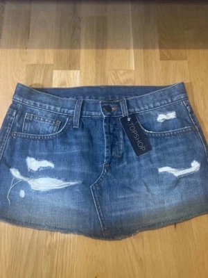 Mini jeanskjol - Helt nya!! Säljer en blå jeanskjol från Topshop med slitna detaljer och rå kant. Kjolen har klassiska fem fickor, bälteshällor och knappstängning framtill. Perfekt för dig som gillar en avslappnad och trendig stil.