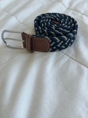 Blått flätat bälte med läderdetaljer - Snyggt flätat bälte i marinblått, grått och vitt med bruna läderdetaljer och silverfärgat spänne. Perfekt accessoar för att lyfta din outfit med en cool och avslappnad vibe.
