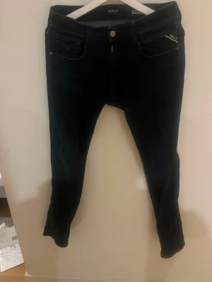 Mörkblå slim fit jeans Replay - Snygga mörkblå slim fit jeans från Replay, modell Anbass. Klassisk femficksdesign med dragkedja och knapp. Jeansen har en smal passform och är tillverkade i stretchigt denimtyg för extra komfort. Perfekta för en trendig och avslappnad stil. Storlek 29/30.