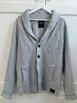 Grå kofta från Hollister - Mysig grå ribbstickad kofta från Hollister med bred sjalkrage och två framfickor. Stängs med svarta knappar framtill och har långa ärmar. Perfekt för lager på lager och chill stil.