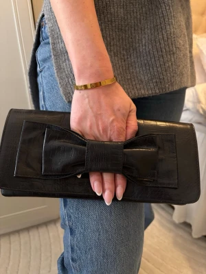 Svart kuvertväska med rosett - Snygg svart kuvertväska i glansigt läderimitation med croco-mönster och stor dekorativ rosett på framsidan. Väskan har ett innerfack med dragkedja och stängs smidigt upptill. Perfekt accessoar för att lyfta din outfit.