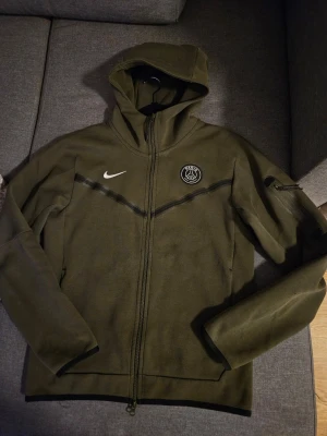 Olivgrön Nike PSG hoodie med dragkedja - ​Säljer min snygga och välvårdade Nike Tech Fleece, bekvämt och varmt material med bra passform.  ​Storlek: S ​Färg: Olivgrön  ​Skick: Mycket fint skick.