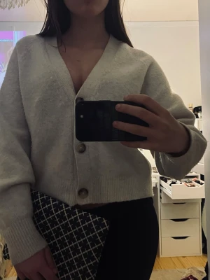 Vit kofta med stora knappar från UNIQLO - Mysig vit kofta med v-ringning och stora dekorativa knappar framtill. Koftan har ribbade muddar vid ärmslut och nederkant samt en avslappnad passform. Perfekt för lager på lager och enkel att styla till olika outfits.