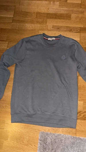 Grå Moncler sweatshirt  - Snygg grå sweatshirt från Moncler i storlek S. Tröjan har rund hals, långärmade ärmar och ett diskret Moncler-logomärke på bröstet. Materialet är mjuk bomull med en lätt textur som ger en clean och modern vibe.