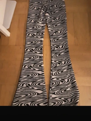Svarta och vita mönstrade utsvängda leggings - Säljer ett par leggings med svartvitt psykedeliskt mönster och utsvängda ben. De har hög midja och är tillverkade i ett stretchigt material som sitter tajt upptill och blir vidare nedtill. Perfekta för dig som vill sticka ut med en cool retrostil.