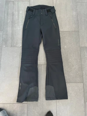 Svarta täckbyxor från Tenson - Snabb leverans, kan lämna till leverans snabbt! Snygga svarta skidbyxor, softshell, från Tenson med dragkedjeförsedda fickor framtill, förstärkta benslut och bälteshällor. Byxorna har en smal passform och är tillverkade i ett slitstarkt, vattenavvisande material som passar perfekt för vintersport.