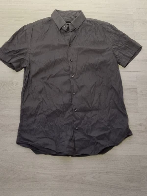 Grå kortärmad skjorta Emporio Armani - Stilren grå skjorta från Emporio Armani med korta ärmar och klassisk krage. Skjortan har knappar framtill och är tillverkad i ett mjukt material som känns skönt mot huden. Perfekt för en clean och enkel look.