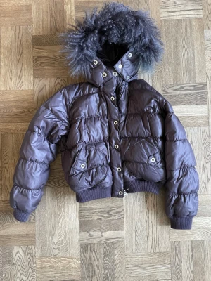 Lila pufferjacka med fuskpäls från Moves - Snygg dovt mörklila pufferjacka från Moves (ser i vissa ljus nästan varmt brun ut) med stor luva och fluffig mjuk fuskpäls. Mycket fint skick! Uppskattas till stl 36-38, men vet ej exakt, känns som S/M. Jackan har breda muddar vid ärmslut och midja, samt stora fickor, dragkedja och med knappstängning framtill. Perfekt för kalla vinterdagar.
