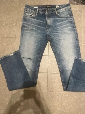Blå slitna jeans från Jack & Jones - Säljer ett par blå jeans från Jack & Jones med slitningar och hål på vänster lår. Jeansen har klassisk femficksmodell, normal midja och dragkedjegylf. Materialet är denim i bomull med en tvättad look och ljusa partier framtill. 31/34