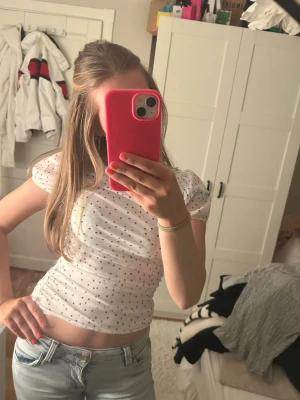 Vit prickig blus med puffärm - Söt vit blus med små svarta prickar och puffiga korta ärmar. Toppen har en figurnära passform och är perfekt för en trendig och ungdomlig look. Passar till både jeans och kjol för en cool stil.