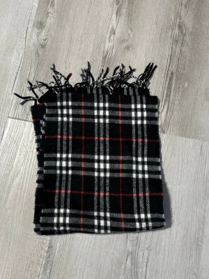 Rutig halsduk från burberry - Snygg halsduk i svart, vitt och rött rutmönster. Halsduken har klassiska fransar i ändarna och är tillverkad i ett mjukt tyg som känns skönt mot huden. Perfekt accessoar för att lyfta din outfit med en tidlös look.